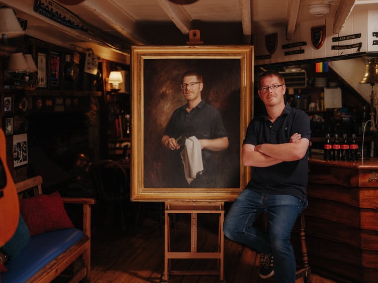 Landlord Simon Dafford-Jones sits next to the portrait of himself painted by Jamie Coreth
